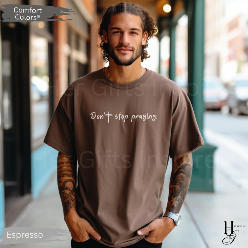 May include: Espresso-colored t-shirt with the text "Don't stop praying." The shirt is a Comfort Colors brand. The model is wearing the shirt outdoors.