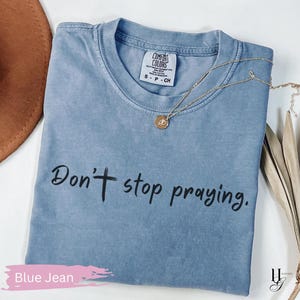 May include: A blue jean colored t-shirt with the text "Don't stop praying" in black script. A gold necklace with a pendant rests on the shirt. The shirt has a Comfort Colors tag.
