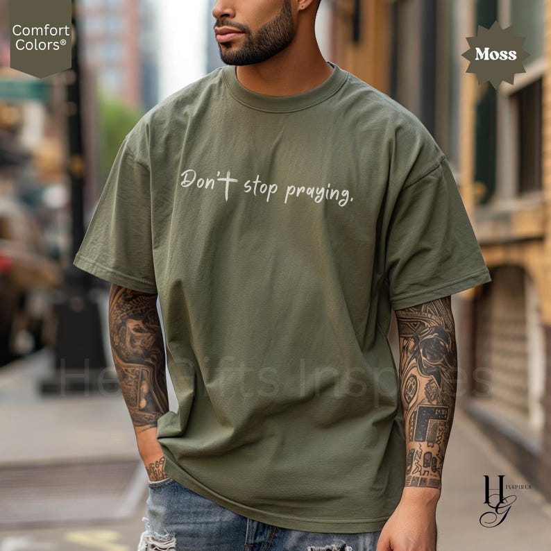 May include: Moss green Comfort Colors t-shirt with the phrase "Don't stop praying" in white script. The shirt is oversized and has short sleeves. The model is wearing blue jeans. The shirt is from the "He Gifts Inspire" collection.