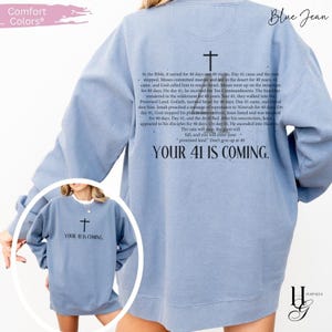 May include: A blue jean colored crewneck sweatshirt featuring a black cross graphic and text on the back detailing biblical events related to the number 40, with the phrase "YOUR 41 IS COMING." A smaller version of the graphic is on the front chest.