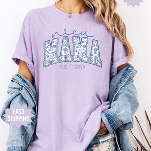 May include: A light purple t-shirt with the text "NICU MAMA" in blue floral print lettering. The shirt also has the text "EST. 2026" in a smaller font. The shirt is a Comfort Colors brand and has a relaxed fit.