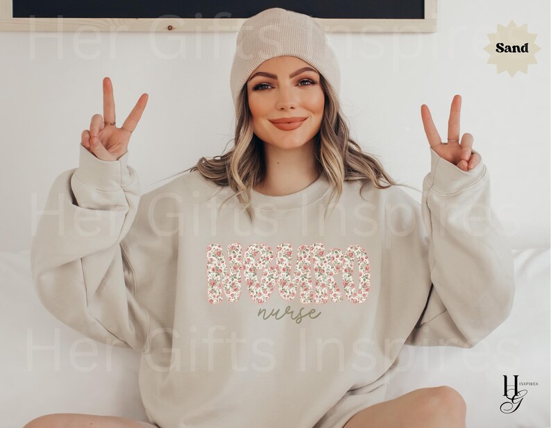 Puede incluir: Sudadera color arena con la palabra "NURSE" en un estampado floral en el pecho. Debajo, la palabra "nurse" est&aacute; escrita en cursiva. La modelo lleva un gorro.