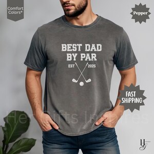 Peut inclure: Un t-shirt gris avec l'inscription blanche "BEST DAD BY PAR", des clubs de golf crois&eacute;s et une balle de golf. Le t-shirt affiche &eacute;galement "EST 2025". Le t-shirt est de la marque Comfort Colors.