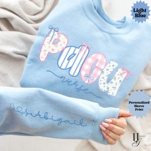 May include: Pale blue sweatshirt featuring the text "PACU nurse" in a decorative font. The letters are adorned with floral, striped, and gingham patterns. The sleeve displays a personalized print with a heartbeat and the name "Abigail".