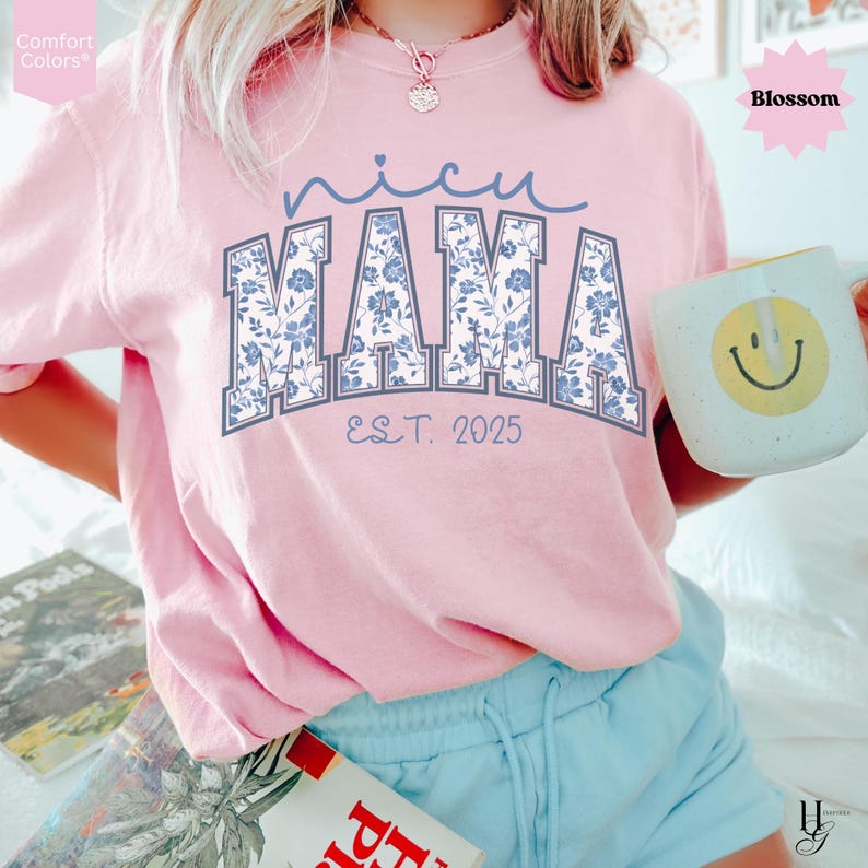 May include: A light pink t-shirt with the words "nicu MAMA" in blue floral print lettering. The text "EST. 2025" is below the word "MAMA". The shirt is a Comfort Colors brand. A white mug with a yellow smiley face is held in the hand.