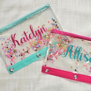 May include: Two clear pencil pouches with zipper closures and metal grommets. One pouch features a teal trim and the name Katelyn in pink glitter script. The other pouch has a hot pink trim and the name Allison in teal script. Both contain colorful star and heart-shaped confetti.
