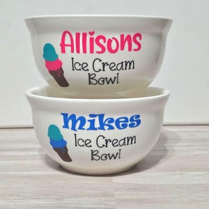 May include: Two white ceramic ice cream bowls stacked on top of each other. The top bowl has the name "Allisons" in pink, with "Ice Cream Bowl" below it. The bottom bowl has the name "Mikes" in blue, with "Ice Cream Bowl" below it. Each bowl has an ice cream cone graphic.