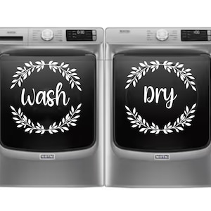 May include: A set of two silver washing machines with a white wreath design on the front. The left machine has the word "wash" inside the wreath, and the right machine has the word "dry" inside the wreath.