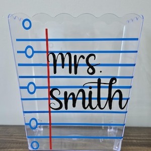 May include: A clear plastic container with a scalloped edge and blue lines resembling notebook paper. The container has a red line down the center and the text "mrs. smith" in black cursive lettering.