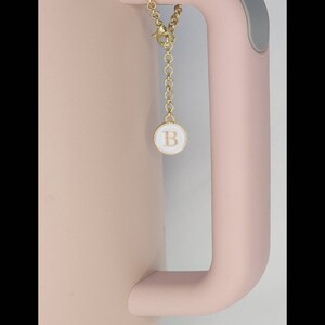 May include: A pink travel mug with a gold chain attached to the handle. The chain has a small round charm with the letter "B" in gold lettering on a white background.