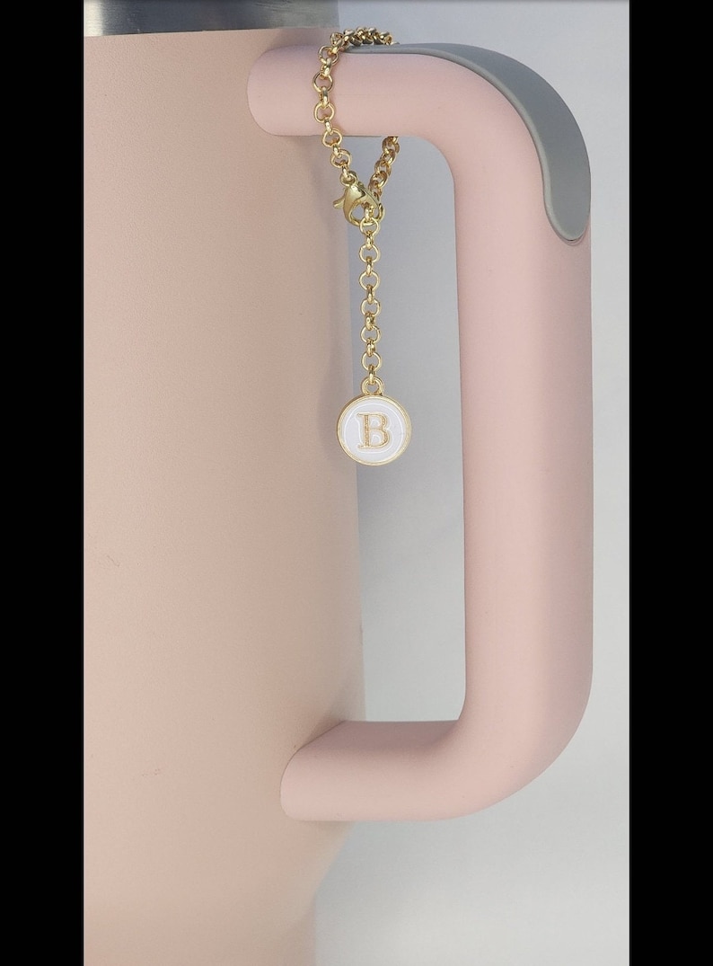 May include: A pink travel mug with a gold chain attached to the handle. The chain has a small round charm with the letter "B" in gold lettering on a white background.