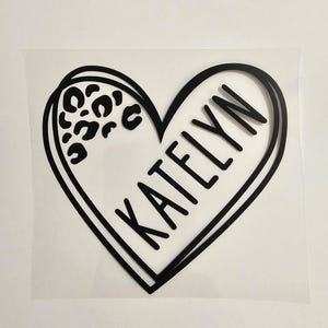 May include: A black vinyl decal featuring a heart shape with a double-line outline. The left side of the heart contains a black leopard print pattern, while the right side displays the name KATELYN in bold, uppercase, sans-serif black lettering.