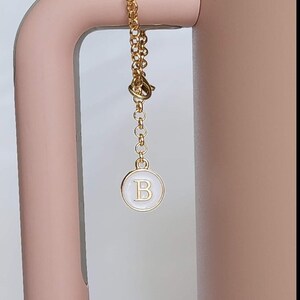 May include: A gold chain with a white circle charm that has the letter "B" in gold lettering.