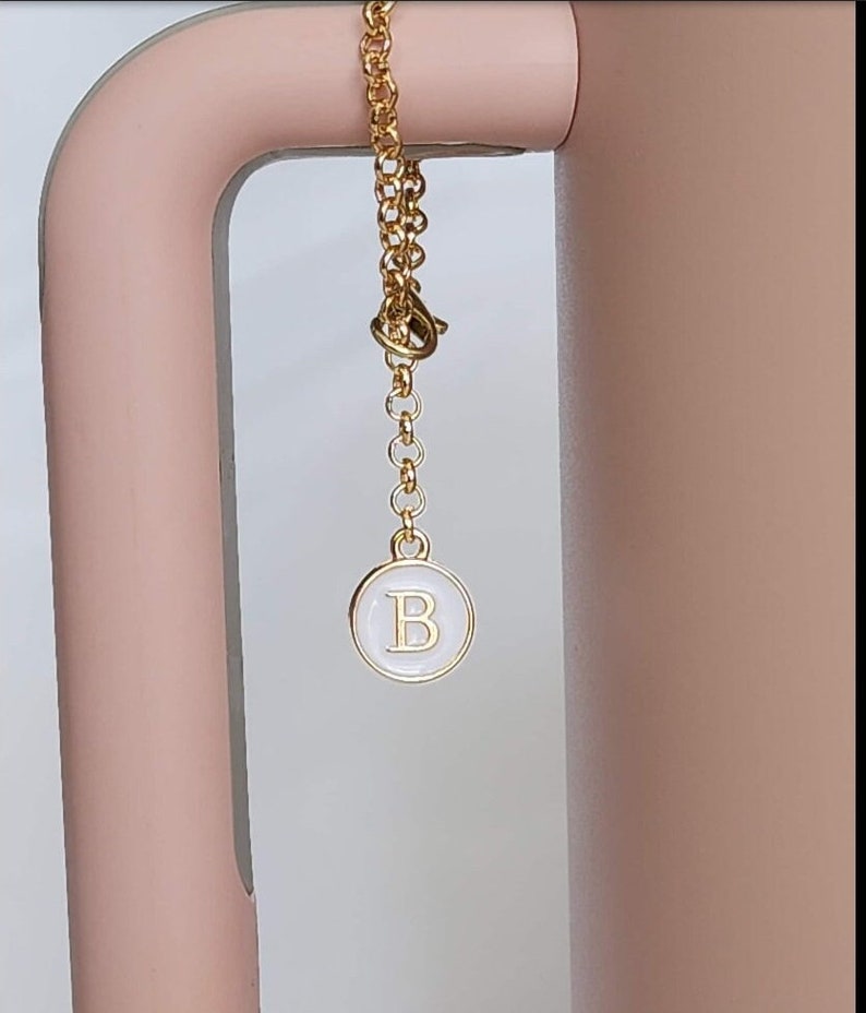 May include: A gold chain with a white circle charm that has the letter "B" in gold lettering.