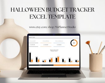 May include: A laptop computer displaying a Halloween budget tracker spreadsheet with a pie chart and bar graphs. The spreadsheet includes categories for costumes, decorations, candy, food, and drinks. The laptop is on a white desk with a vase of paintbrushes.
