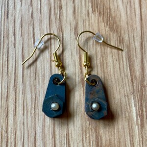 Könnte beinhalten: Ein Paar Ohrringe mit einem goldenen Haken und einer kleinen, runden, weißen Perle. Die Ohrringe sind aus Metall und haben einen rustikalen, industriellen Look.
