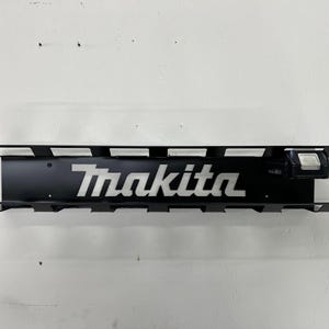 May include: Black metal Makita tool holder with the Makita logo in white. The holder has slots for tools and a battery. The holder is mounted on a white wall.