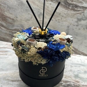 May include: A black round box with a blue and white dried flower arrangement. The box has a gold logo that says "Aneka's Collection". A black diffuser stick set is in the centre of the arrangement.