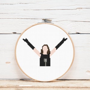 May include: A cross-stitch embroidery hoop with a black and white pixelated image of a person with brown hair wearing a black dress and black gloves, with arms raised in the air.