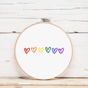 Peut inclure: Un cercle de broderie au point de croix avec six cœurs pixélisés aux couleurs de l'arc-en-ciel : rouge, orange, jaune, vert, bleu et violet. La broderie est réalisée sur un fond de tissu blanc, maintenue dans un cercle en bois. Le fond est une surface en bois blanc.