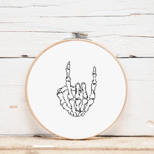 May include: Black and white cross-stitch embroidery of a skeleton hand making the rock and roll hand gesture. The embroidery is in a round wooden embroidery hoop.