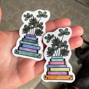 May include: Two stickers featuring a stack of books with a plant growing out of the top. The books are in shades of blue, purple, green, and yellow. The plants are green with a light green stem. The stickers are on a hand.