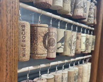 May include: A close-up of a wall display of cork earrings with various designs and text, including "Wine Rocks", "Moscato d'Asti", "Galliano", "Friendliestates.com", and "100% Pro-Mas".