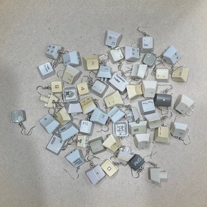 Puede incluir: Pendientes hechos a mano con teclas de teclado de ordenador recicladas. Los pendientes presentan teclas en tonos grises, beige y blanco, con ganchos plateados. Las teclas muestran letras, números y símbolos.