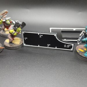 May include: A black measuring tool with white markings and measurements in inches, ranging from 0.5 to 3 inches. Miniature figures of green and blue soldiers are in the background, suggesting a tabletop game.