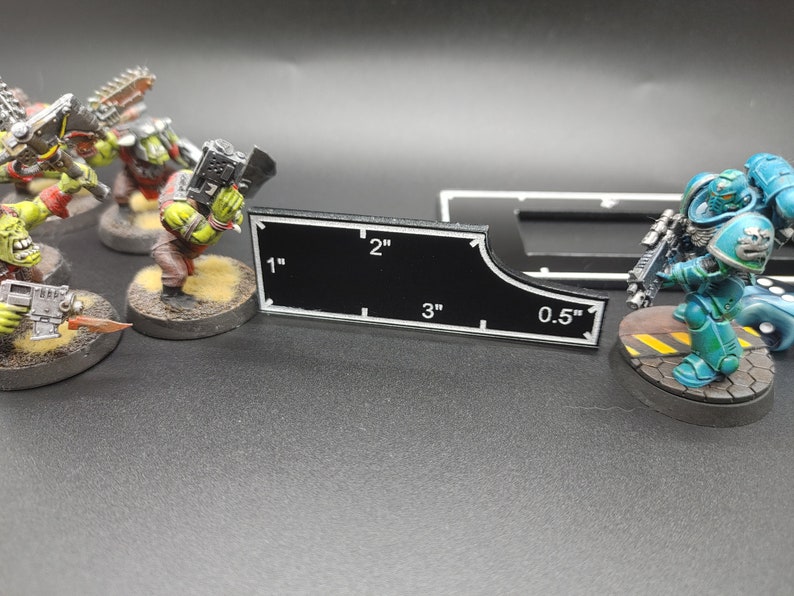 May include: A black measuring tool with white markings and measurements in inches, ranging from 0.5 to 3 inches. Miniature figures of green and blue soldiers are in the background, suggesting a tabletop game.