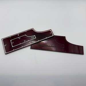 May include: Two burgundy acrylic quilting rulers with white markings. One ruler has a rectangular shape with a cutout and measurements in inches. The other ruler is a smaller, curved shape with inch markings. Ideal for quilting projects.