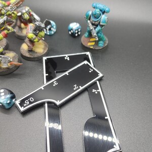 May include: A tabletop gaming scene featuring miniature figures of orcs and a space marine, dice, and black measuring tools with white markings in inches. The scene is set on a dark surface.