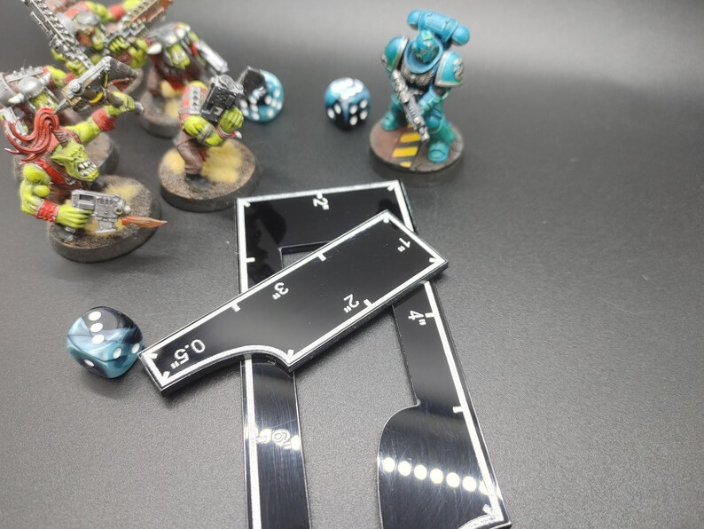 May include: A tabletop gaming scene featuring miniature figures of orcs and a space marine, dice, and black measuring tools with white markings in inches. The scene is set on a dark surface.