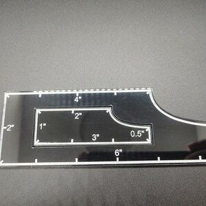 May include: Black acrylic quilting ruler with white markings. Features measurements in inches, including 6" and 0.5" increments. The ruler has a unique shape with a curved edge and a rectangular cutout.