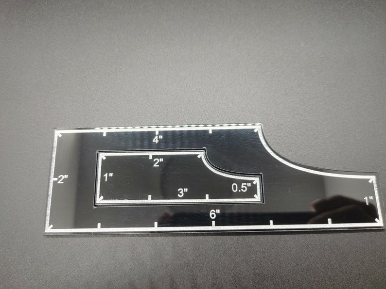 May include: Black acrylic quilting ruler with white markings. Features measurements in inches, including 6" and 0.5" increments. The ruler has a unique shape with a curved edge and a rectangular cutout.