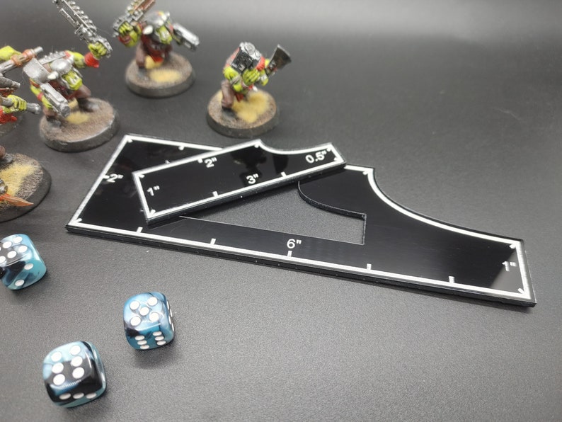 May include: Black acrylic gaming templates with white markings, including measurements of 0.5", 1", 2", 3", and 6". Miniature figurines and dice are also present, suggesting a tabletop gaming theme.