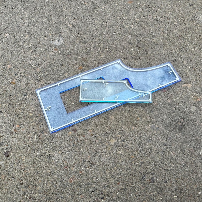 May include: Three clear acrylic quilting rulers with blue edges are displayed on a gray surface. The rulers include a rectangular shape with a cut-out, a curved shape, and a smaller rectangular ruler. Measurements are in inches.