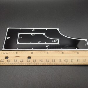 May include: A black acrylic quilting template with white markings and measurements in inches. The template has a unique shape with a rectangular cutout and curved edges. A wooden ruler is placed next to the template for scale.