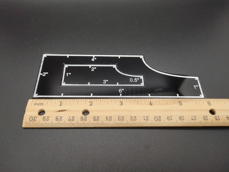 May include: A black acrylic quilting template with white markings and measurements in inches. The template has a unique shape with a rectangular cutout and curved edges. A wooden ruler is placed next to the template for scale.