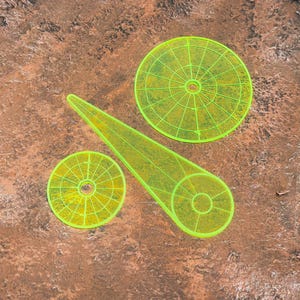 May include: A set of three neon yellow acrylic templates for quilting or sewing, consisting of one large circular grid, one smaller circular grid, and one elongated wedge-shaped piece, all featuring precise etched markings for alignment.