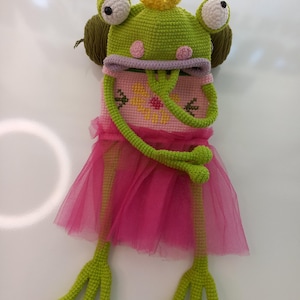 May include: A whimsical, handmade frog costume with a green crocheted head, large white eyes, and a yellow crown. The costume includes a pink cross-stitched bodice and a pink tulle skirt, with long green crocheted arms and legs.