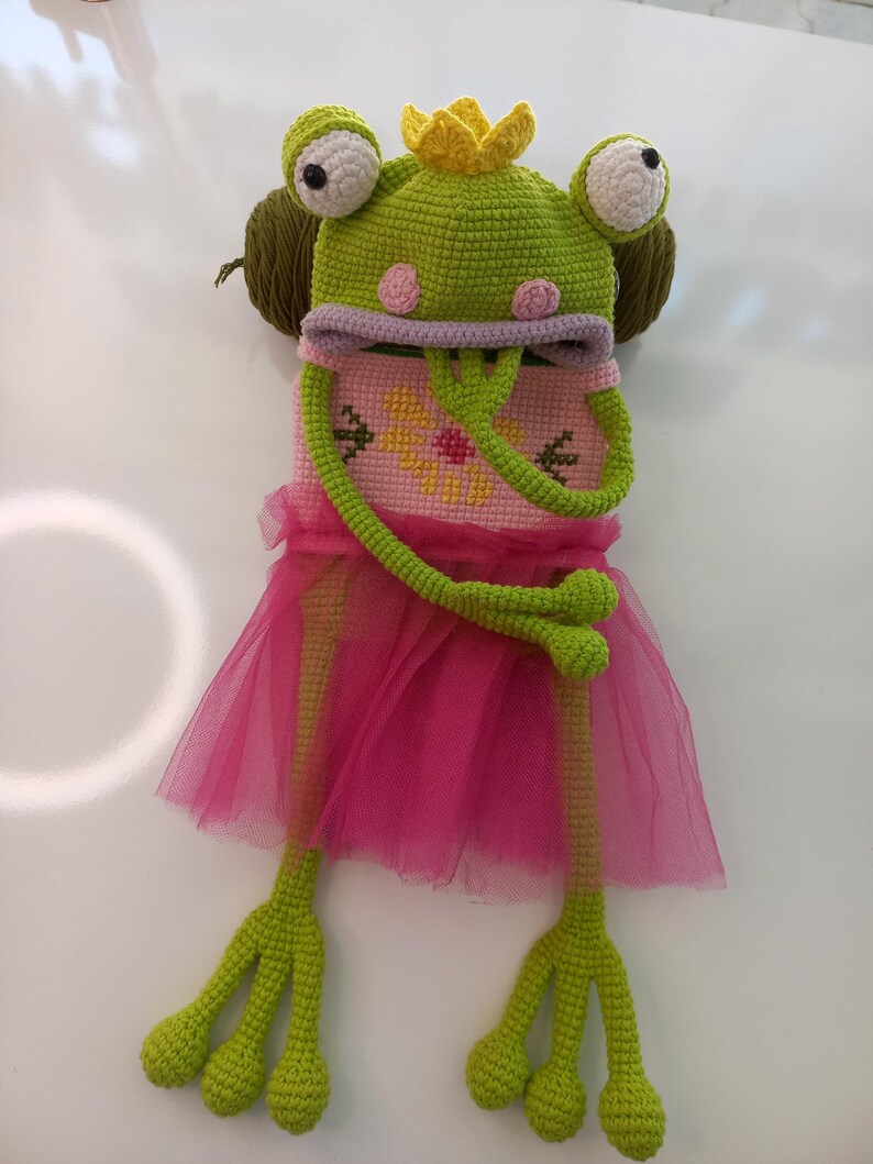 May include: A whimsical, handmade frog costume with a green crocheted head, large white eyes, and a yellow crown. The costume includes a pink cross-stitched bodice and a pink tulle skirt, with long green crocheted arms and legs.