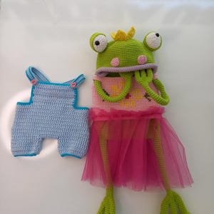 May include: Crocheted frog toy wearing a pink tutu and a yellow crown. The frog has large, round eyes and long, green legs. Next to it is a blue crocheted overall.