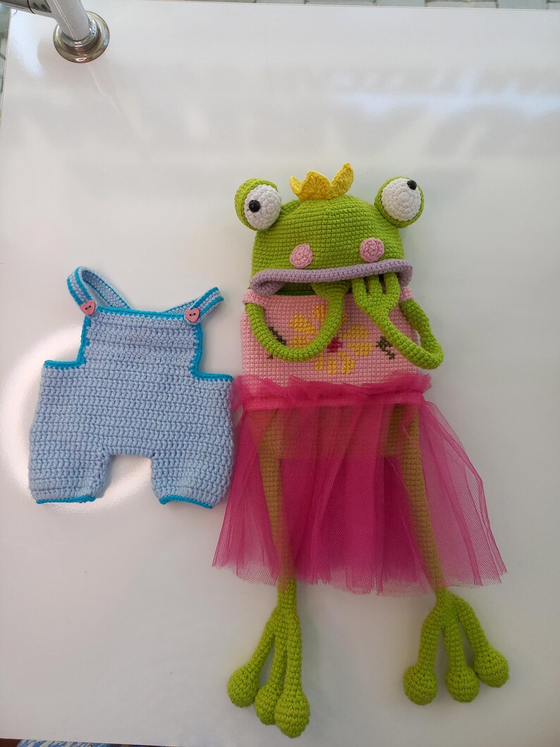 May include: Crocheted frog toy wearing a pink tutu and a yellow crown. The frog has large, round eyes and long, green legs. Next to it is a blue crocheted overall.