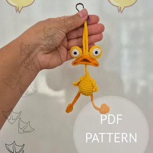 Puede incluir: Un llavero de pato amarillo hecho a mano con crochet, con grandes ojos saltones y patas naranjas. El pato está suspendido de un anillo de metal. El texto "PDF PATTERN" es visible en un círculo blanco.