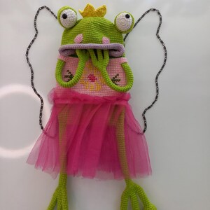 May include: A whimsical crochet frog purse with a green frog hat, large white-eyed details, and a yellow crown. The frog wears a pink dress with a tulle skirt and has long green legs. A silver chain is attached for carrying.