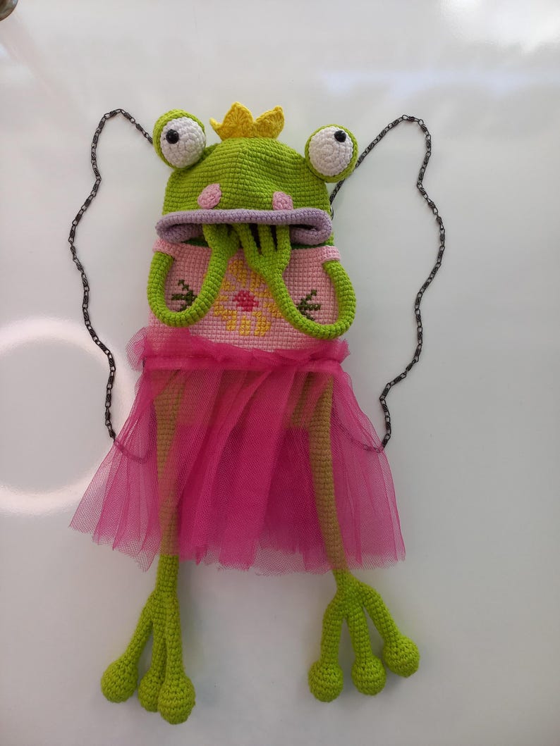 May include: A whimsical crochet frog purse with a green frog hat, large white-eyed details, and a yellow crown. The frog wears a pink dress with a tulle skirt and has long green legs. A silver chain is attached for carrying.