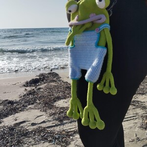 Peut inclure: Une grenouille verte au crochet faite &agrave; la main, avec de grands yeux et une longue langue rose, portant une salopette bleu clair avec une bordure blanche. La grenouille est suspendue &agrave; une cha&icirc;ne, avec une plage et l'oc&eacute;an en arri&egrave;re-plan.