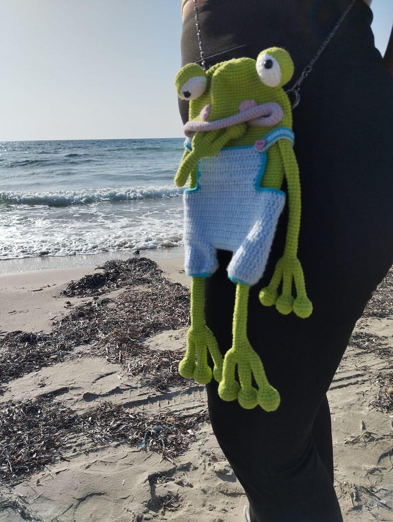 Peut inclure: Une grenouille verte au crochet faite &agrave; la main, avec de grands yeux et une longue langue rose, portant une salopette bleu clair avec une bordure blanche. La grenouille est suspendue &agrave; une cha&icirc;ne, avec une plage et l'oc&eacute;an en arri&egrave;re-plan.