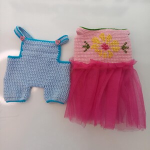 May include: Two crocheted doll outfits. One is a light blue overall with blue trim and pink heart-shaped buttons. The other is a pink dress with a yellow flower design and a pink tulle skirt. The outfits are handmade.